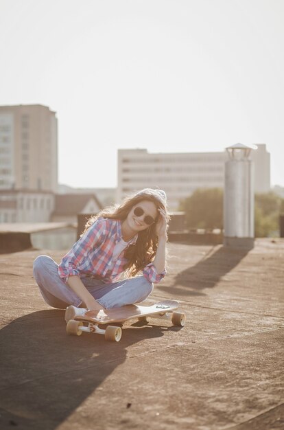 Chica con estilo fresco con patineta sentada en el techo de la ciudad