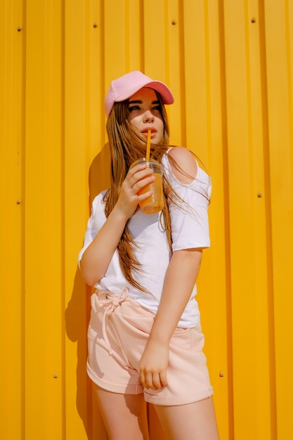 Chica de estilo hipster bastante joven divertirse al aire libre con gorra blanca y comer helado en amarillo