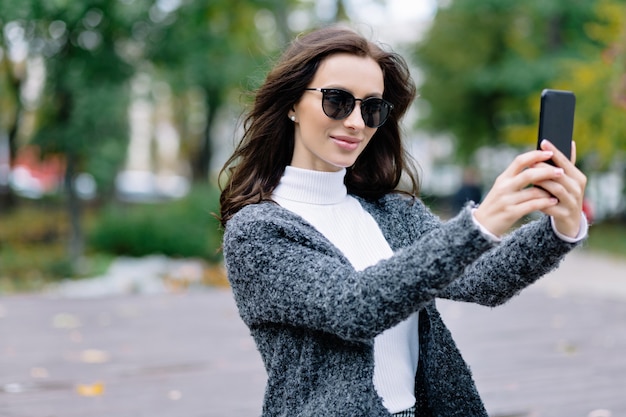 Chica de estilo sonriente con cabello oscuro disfrutando de caminar en el parque y hacer selfie. Retrato al aire libre de la joven mujer riendo en traje de moda tomando una foto de sí misma al lado del hermoso parque de otoño