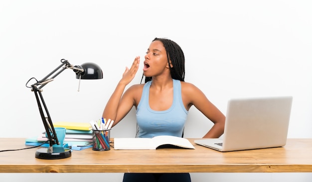 Chica estudiante adolescente afroamericana con cabello largo trenzado en su lugar de trabajo bostezando