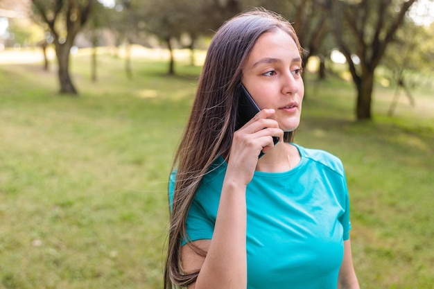 Chica estudiante adolescente con camiseta color aguamarina, haciendo una llamada telefónica a su familia en el parque