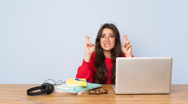 Chica estudiante adolescente estudiando en una mesa con los dedos cruzados y deseando lo mejor