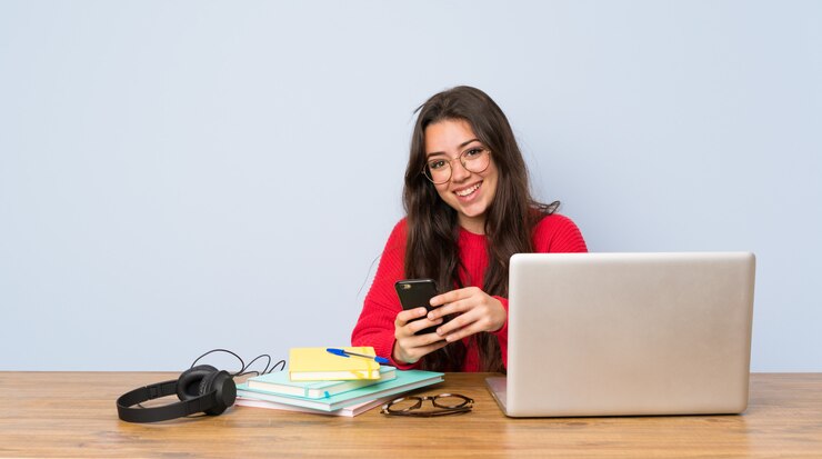 Foto chica estudiante adolescente estudiando en una mesa enviando un mensaje con el móvil