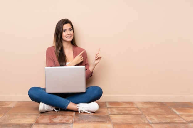 Chica estudiante adolescente sentada en el suelo con una computadora portátil asustada y apuntando hacia un lado
