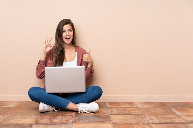 Chica estudiante adolescente sentada en el suelo con un portátil mostrando signo bien y pulgar arriba gesto