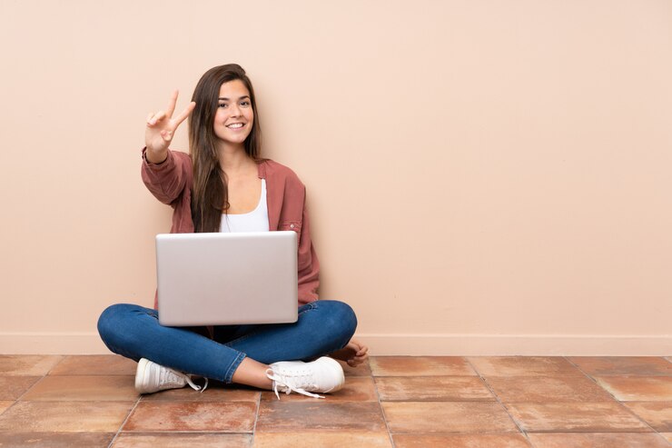 Foto chica estudiante adolescente sentada en el suelo con un portátil sonriendo y mostrando el signo de la victoria
