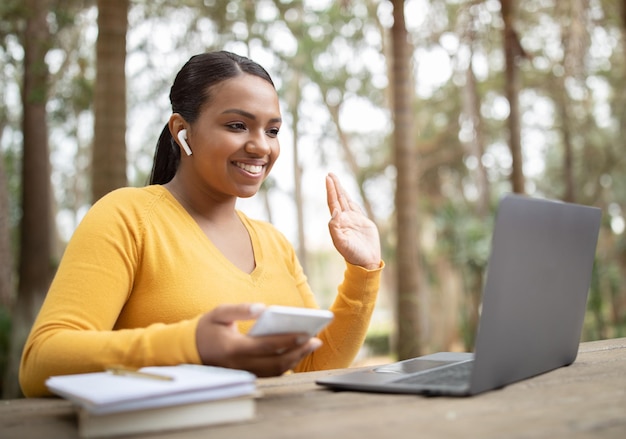 Chica estudiante alegre saludando con la mano a la computadora portátil mientras hace videollamadas aprendiendo en línea sentada en el parque