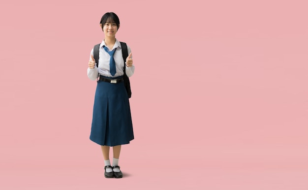 Chica estudiante asiática de cuerpo completo con uniforme con una sonrisa dando pulgares hacia arriba aislada en un fondo rosa con caminos de recorte para el trabajo de diseño espacio libre vacío