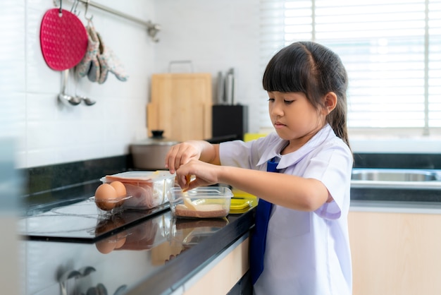 Chica estudiante de escuela primaria en uniforme haciendo sándwich para lonchera en la rutina escolar matutina