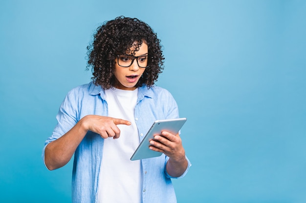 Chica estudiante estadounidense sorprendida con pelo rizado africano sosteniendo tableta digital sobre fondo azul aislado con espacio de copia de texto, logotipo o publicidad.