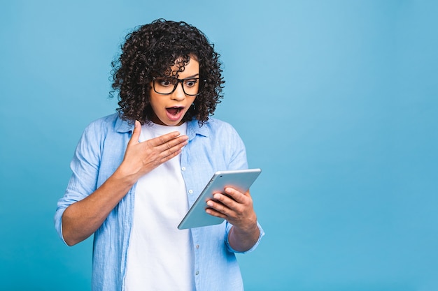 Chica estudiante estadounidense sorprendida con pelo rizado africano sosteniendo tableta digital sobre fondo azul aislado con espacio de copia de texto, logotipo o publicidad.