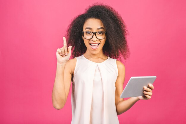 Chica estudiante estadounidense sorprendida con pelo rizado africano sosteniendo tableta digital sobre rosa con espacio de copia de texto, logotipo o publicidad.