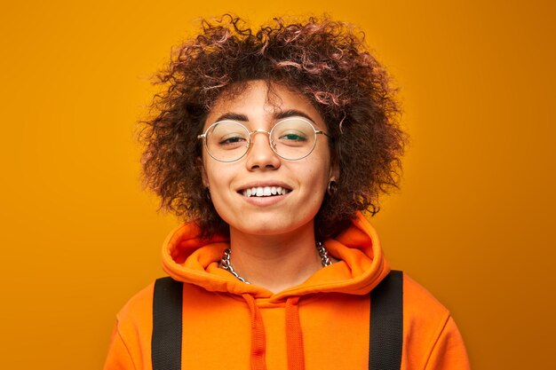 Foto chica estudiante kazaja positiva con peinado afro y mochila sonriendo sobre fondo de estudio amarillo