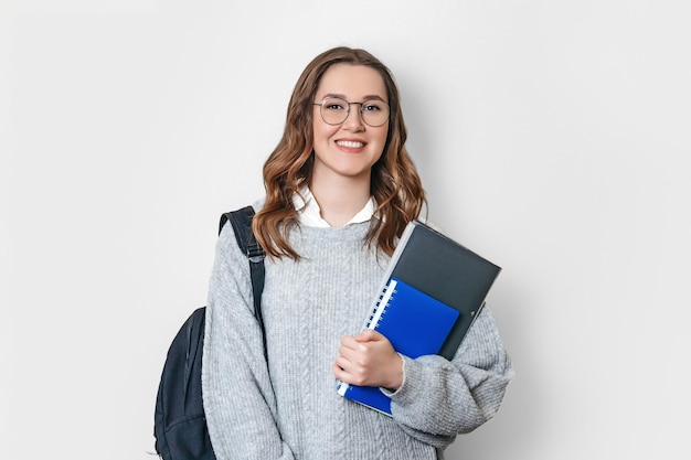 Chica estudiante con mochila sonríe y sostiene una carpeta de cuaderno sobre un fondo blanco