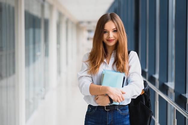 Chica estudiante morena alegre con mochila negra tiene libros en edificio moderno