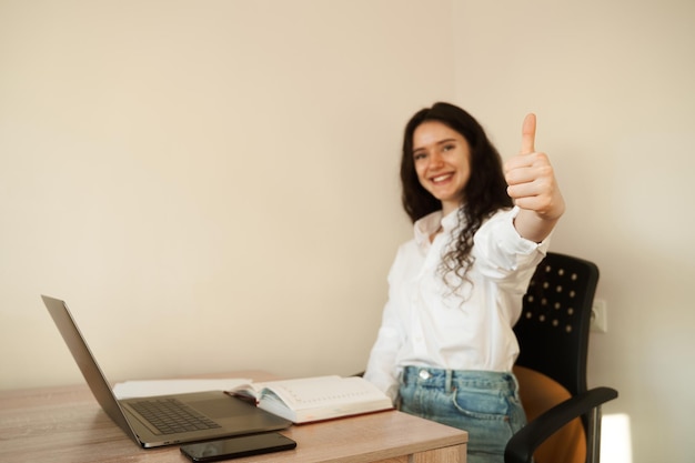 Chica estudiante mostrando los pulgares hacia arriba Colegiala con la computadora portátil hecha la tarea en línea y gesticulando con los pulgares hacia arriba en la mesa en casa