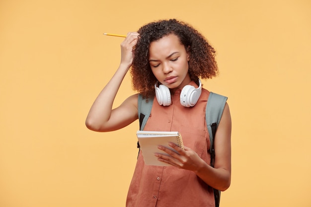 Chica estudiante negra concentrada con auriculares inalámbricos alrededor del cuello rascándose la cabeza mientras piensa en la solución de la tarea