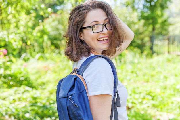 Chica estudiante positiva feliz en anteojos con mochila sonriendo en parque verde