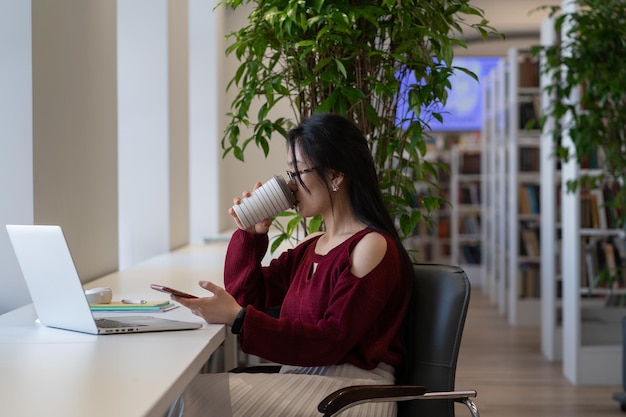 Chica estudiante pospone las cosas en la biblioteca con chat de teléfono inteligente en las redes sociales sin prepararse para el examen