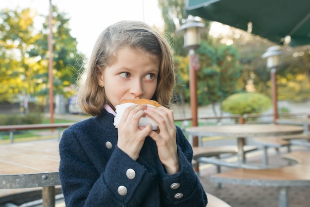 Chica estudiante de primaria come hamburguesa, sandwich en un café al aire libre