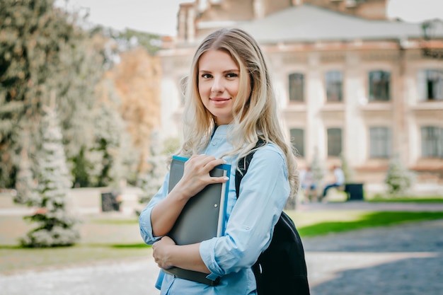 chica estudiante rubia está sonriendo y sosteniendo una carpeta y un cuaderno en sus manos en la universidad