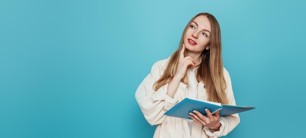 Chica estudiante rubia pensativa sosteniendo un libro de ejercicios abierto y buscando copiar espacio