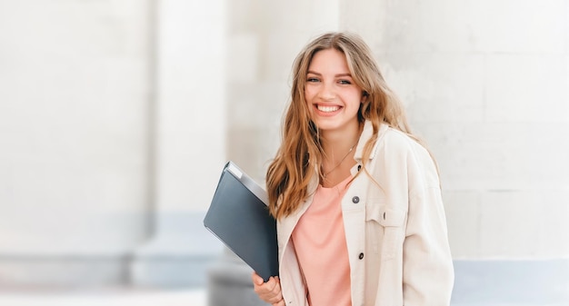 Chica estudiante rubia sonriendo contra la universidad y tiene carpetas y cuadernos en las manos