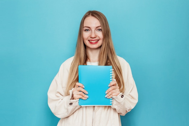 Chica estudiante rubia sonriente sosteniendo un libro, portátil sonriendo, mirando a la cámara aislada en la pared azul