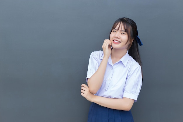 Chica estudiante de secundaria asiática en el uniforme escolar con sonrisas confiadas con gris en el fondo