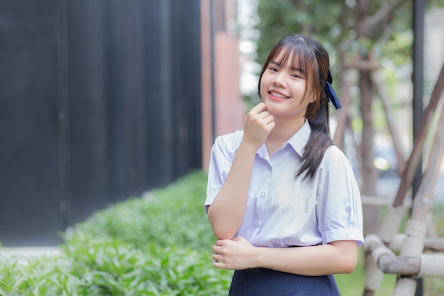 Chica estudiante de secundaria asiática en uniforme escolar con sonrisas confiadas con el jardín del edificio de la ciudad