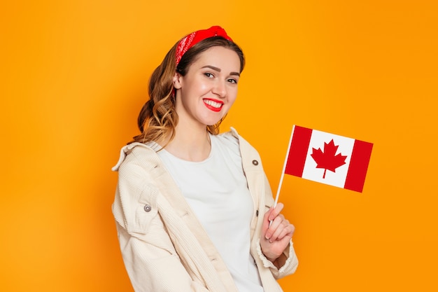 Chica estudiante sonriendo y sosteniendo una pequeña bandera de Canadá aislada sobre fondo naranja