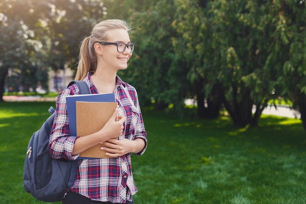 Chica estudiante sonriente feliz con libros en el parque al aire libre, mirando a otro lado, soñando o pensando durante las vacaciones, descansando en el campus. Concepto de educación, espacio de copia