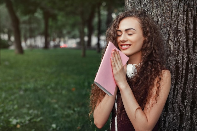 Chica estudiante sosteniendo un libro y sentada bajo un árbol