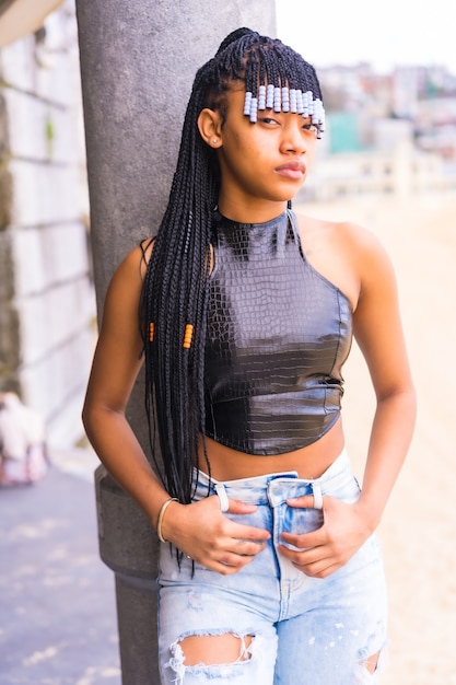 Chica étnica negra con trenzas con top de cuero y pantalón vaquero. Disfrutando de las vacaciones de verano junto a la playa, foto vertical