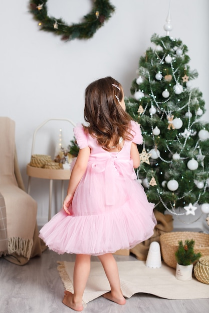 Chica europea con cabello rubio en un elegante vestido rosa cerca de un árbol de Navidad bailando