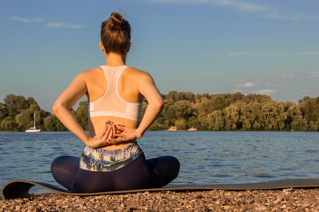 Una chica europea en un chándal ligero practica yoga en la orilla del río sobre una estera de goma para yoga