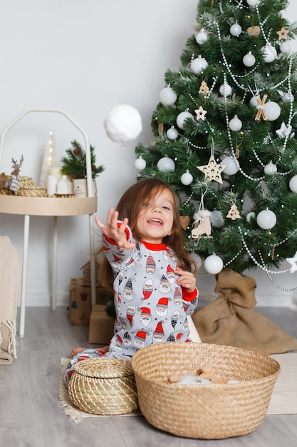 Chica europea en pijama con santa junto al árbol de navidad lanza una bola de nieve a la cámara