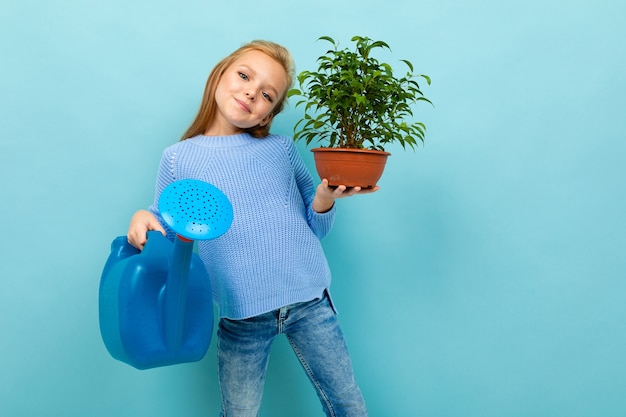 Chica europea con una regadera y una planta en sus manos sobre una pared azul claro.