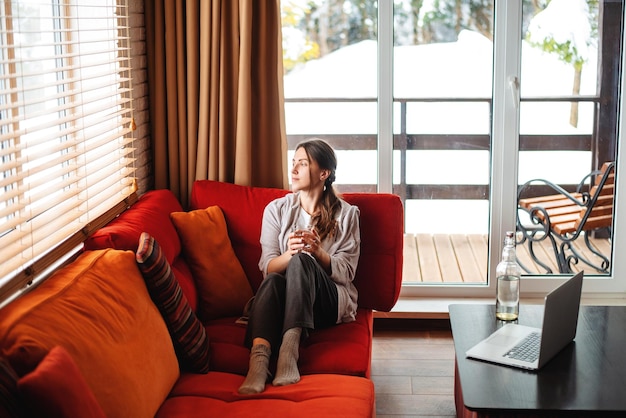 Chica europea sentada en el sofá y bebiendo agua de un vaso en casa Mujer joven pensativa usa ropa informal Interior de la sala de estar en un apartamento moderno Computadora portátil y botella en la mesa