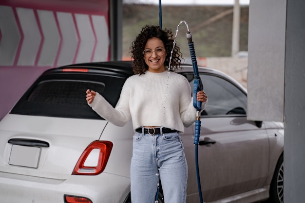 Chica europea sostenga la manguera de agua en el lavado de autos cerca del automóvil personal. Hermosa joven sonriente rizada con gafas y mirando a la cámara