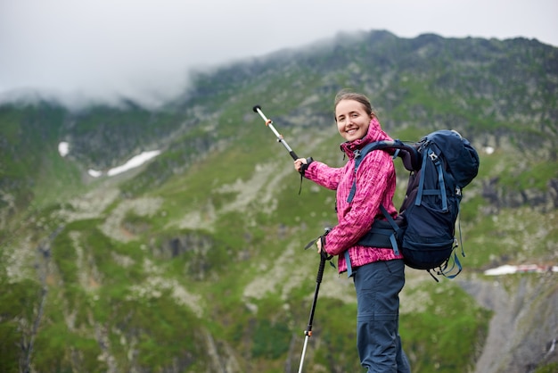 Chica excursionista apuntando con un palo de trekking en la cresta verde de las montañas rocosas con nubes brumosas