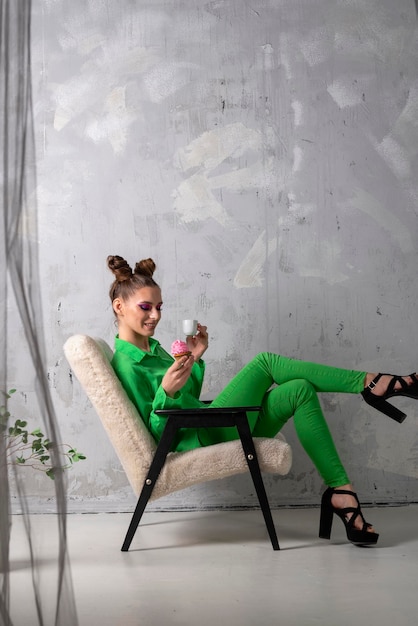 Chica extraordinaria en traje verde con taza de café y muffin Modelo con maquillaje brillante sobre fondo gris