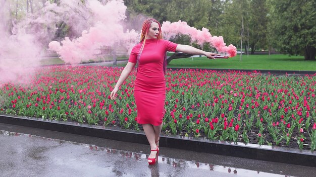 Una chica extraordinaria con un vestido rojo con maquillaje y trenzas de colores. Lindo sonriendo y posando en un espeso humo rosa en el parque bajo la lluvia