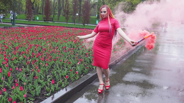 Una chica extraordinaria con un vestido rojo con maquillaje y trenzas de colores. Lindo sonriendo y posando en un espeso humo rosa en el parque bajo la lluvia