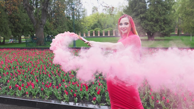 Una chica extraordinaria con un vestido rojo con maquillaje y trenzas de colores. Lindo sonriendo y posando en un espeso humo rosa en el parque bajo la lluvia