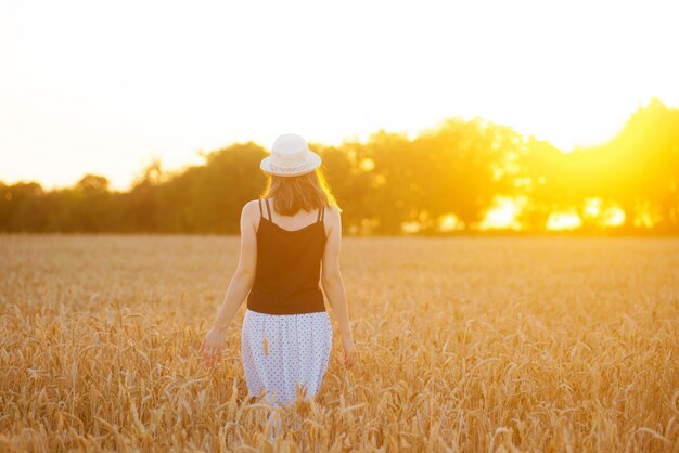 Chica con falda larga y sombrero está caminando en un campo de cultivos en la luz del sol.