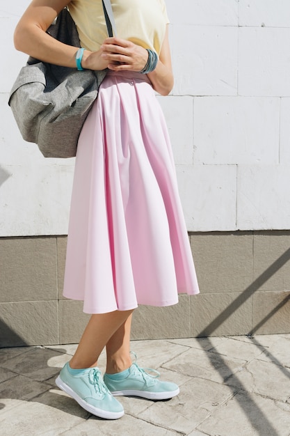 Chica en falda rosa, camisa amarilla y con una mochila de pie contra una pared blanca