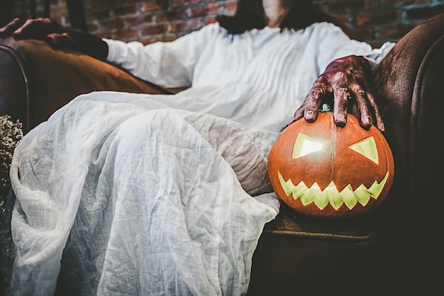 Chica fantasma en la sangre con vestido blanco sosteniendo la calabaza de halloween y sentado en la vieja