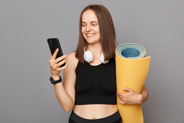 Una chica feliz y alegre sosteniendo una alfombra de fitness en sus manos posando aislada sobre un fondo gris usando un teléfono móvil después de hacer ejercicio publicando una foto en la red social