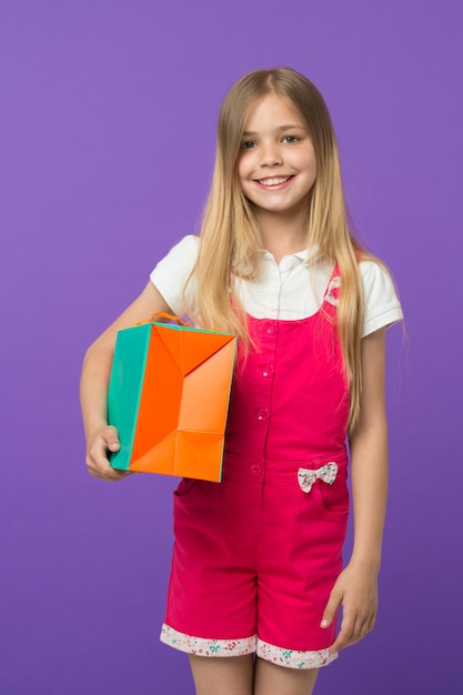 Chica feliz con bolsa de compras sobre fondo violeta. Pequeña sonrisa adicta a las compras con bolsa de papel. Comprador infantil en mono de moda. Preparación y celebración de fiestas. Compras y venta.